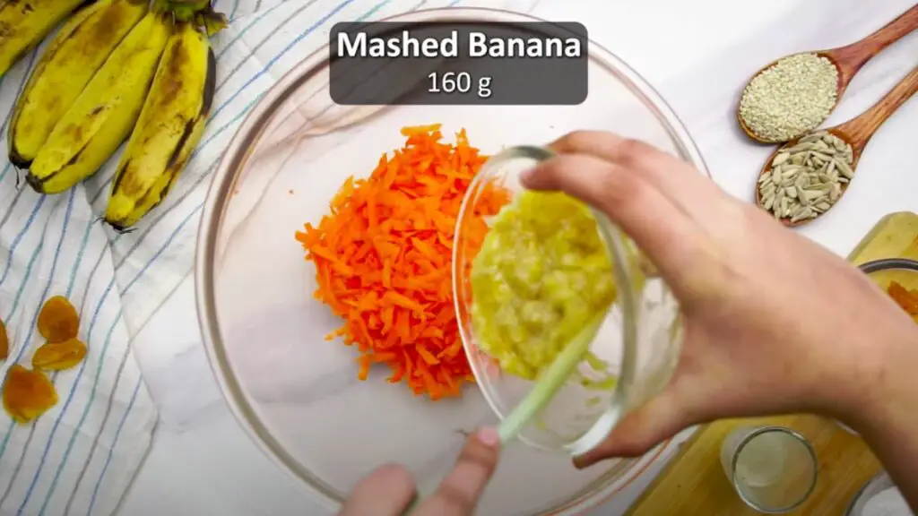 adding mashed bananas to the grated carrots in a mixing bowl