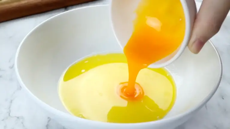 A hand is pouring a vibrant orange egg yolk mixture into a bowl of melted butter on a marble countertop.