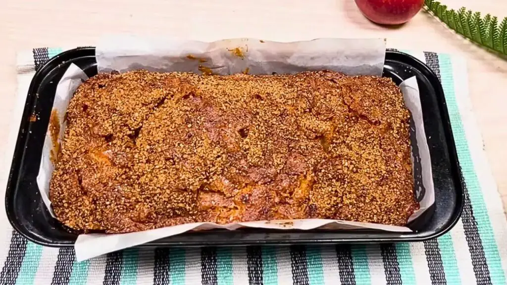 Freshly baked apple cinnamon loaf cake in a parchment-lined loaf pan, topped with a golden cinnamon sugar crust and resting on a striped kitchen towel.