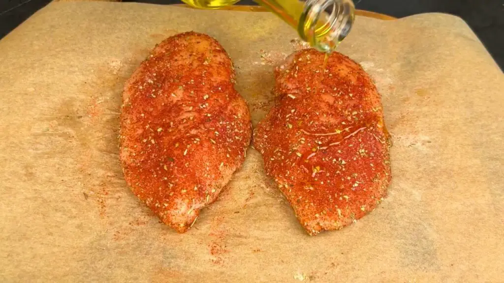 Air fryer chicken breast seasoned with spices on parchment paper as olive oil is drizzled over the top.