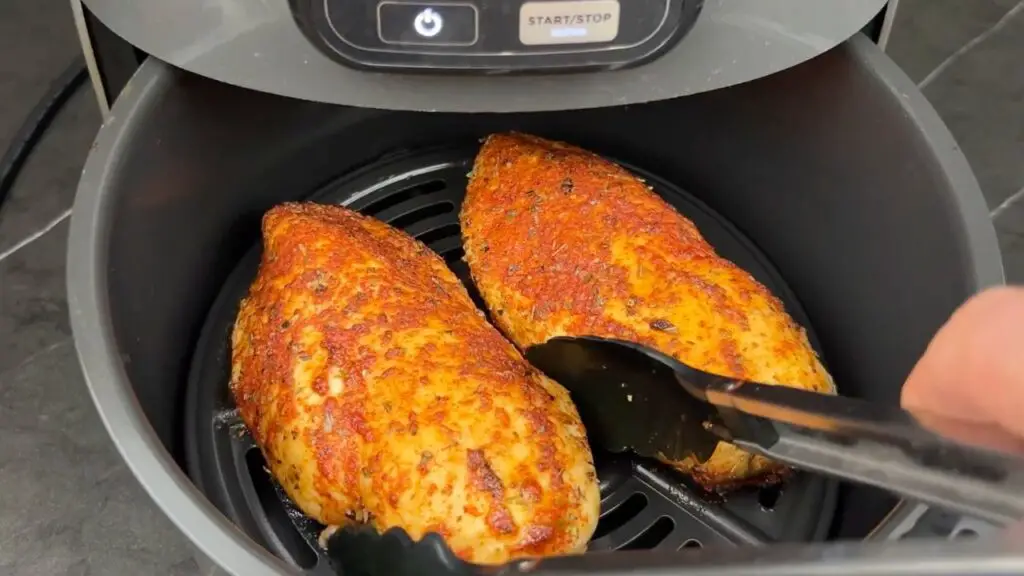 Best air fryer chicken breast cooking in an air fryer basket, showing two golden, seasoned chicken breasts being turned with tongs.