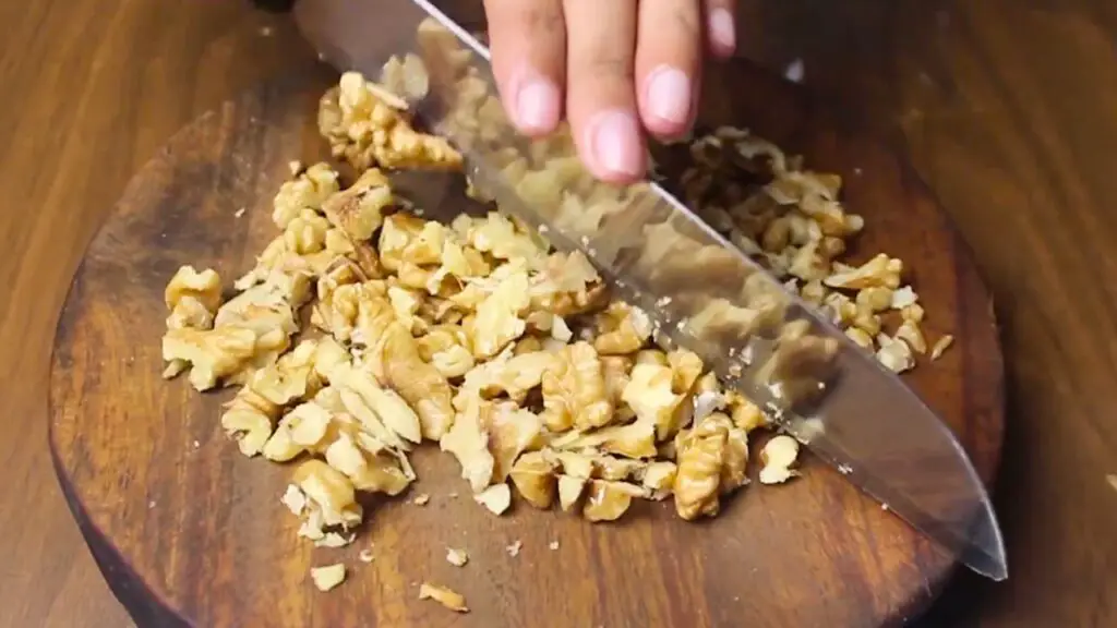 Chopping walnuts on a wooden cutting board with a knife, preparing nuts to be added into the batter for a healthy oat cake recipe.