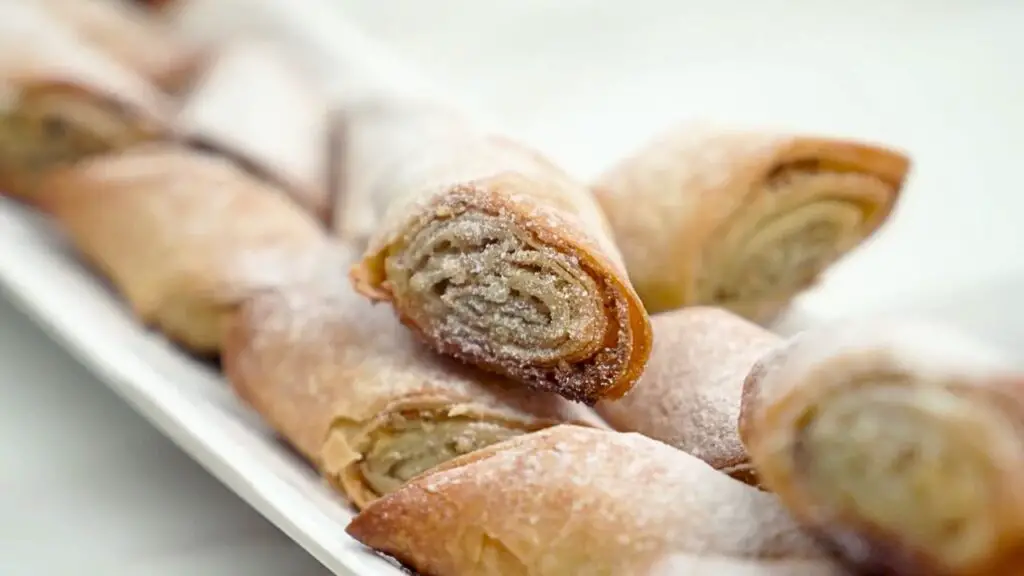 Close-up of golden, flaky baklava rolls with phyllo dough, showing layered pastry and nut filling dusted lightly with sugar
