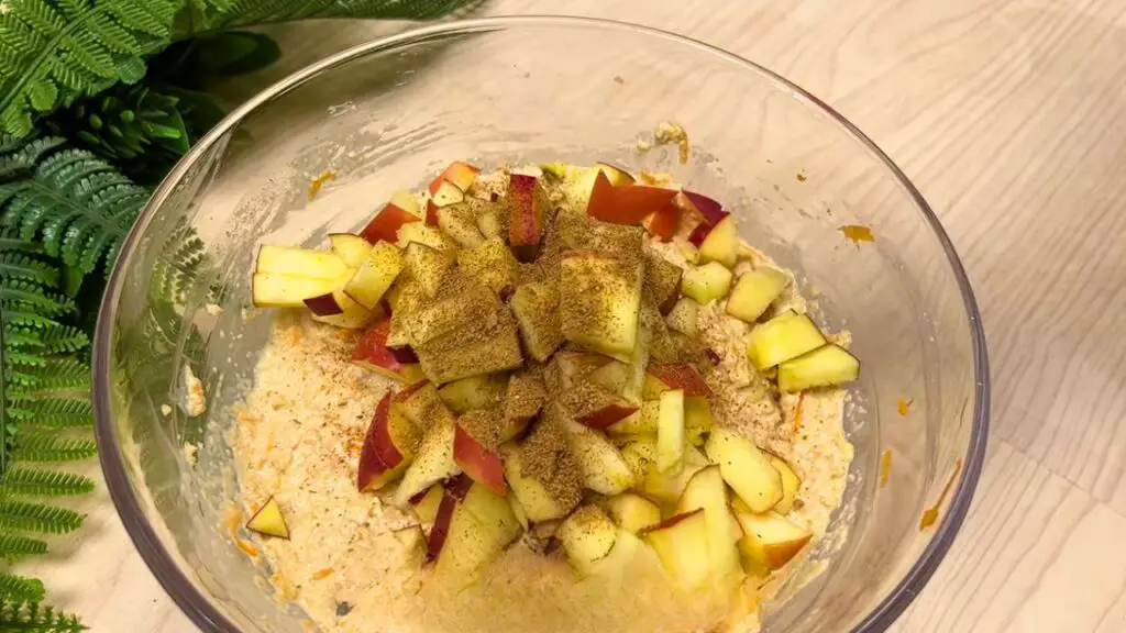 Glass mixing bowl containing oatmeal batter with added apple and cinnamon, prepared for an oatmeal apple and carrot cake