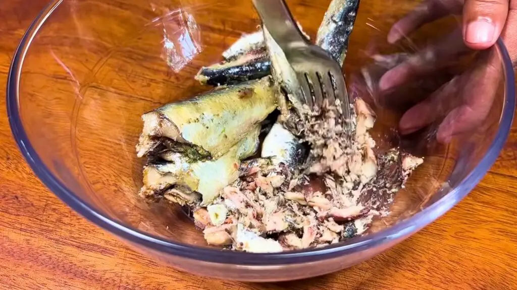  use a fork to break drained sardines into flaky pieces in a clear glass bowl on a wooden surface, preparing the mixture for sardine fish cakes.