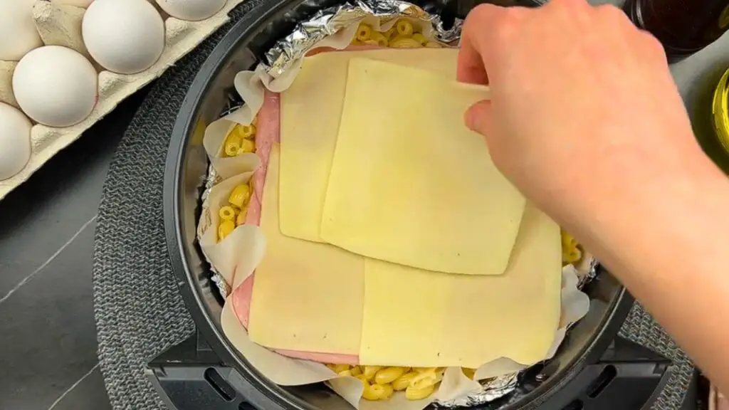 Placing ham and cheese slices over pasta in an air fryer basket for an air fryer pasta recipe.