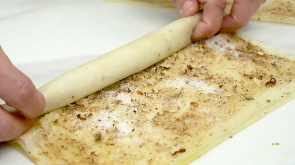Rolling phyllo dough into a tight log, keeping the filling snug to form baklava rolls with phyllo dough