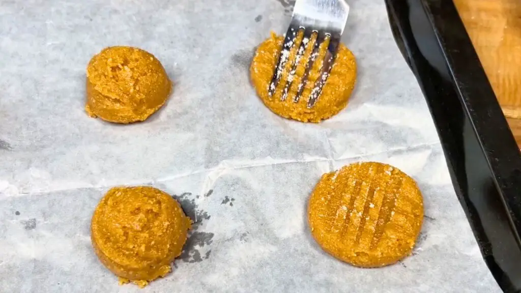 A fork presses down cookie dough balls on a parchment-lined baking sheet to create a crisscross pattern for flourless peanut butter cookies.