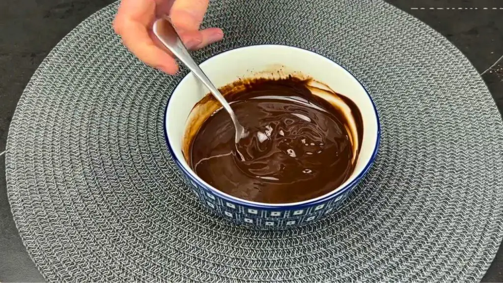A hand stirring melted chocolate and butter in a patterned bowl with a spoon, showing the smooth glossy texture, for how to make individual brownies.