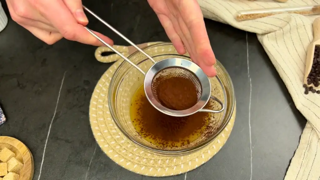 Cocoa powder being sifted into a glass bowl of batter for air fryer chocolate cupcakes.