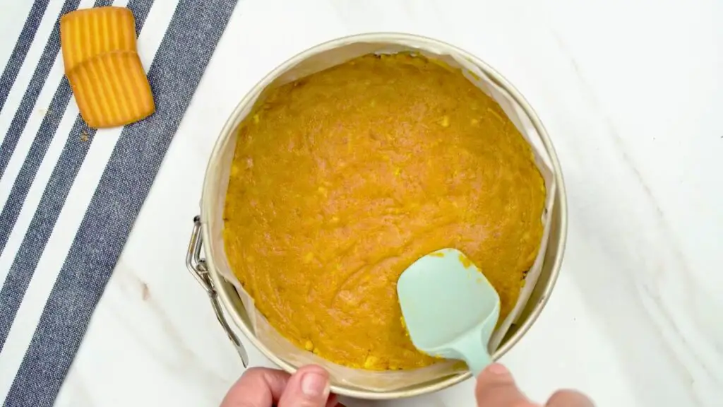 Crust mixture pressed firmly into a lined springform pan with a spatula to form an even base for a no-bake cheesecake.