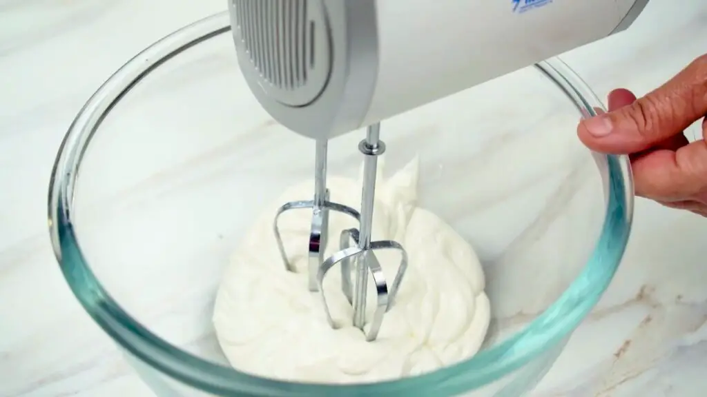 Heavy cream being whipped with an electric mixer until thick, soft peaks form, part of an easy no bake cheesecake recipe.