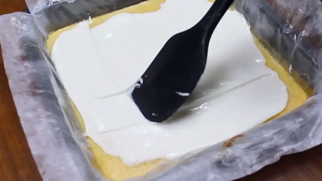 Spreading thick lemon glaze over baked bars for easy lemon brownies in a parchment-lined pan, smoothing the icing evenly across the top.