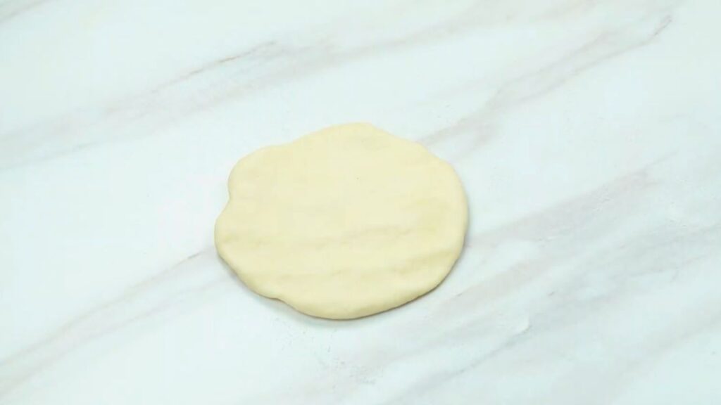Beginner croissant recipe step showing a small, flattened round of dough resting on a clean countertop, ready for the next stage after rising.