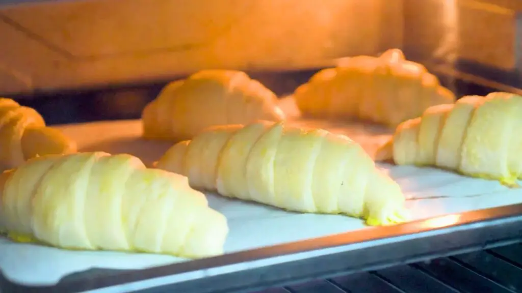 Beginner croissant recipe step showing croissants baking in the oven on a parchment-lined tray, starting to puff up and turn lightly golden.