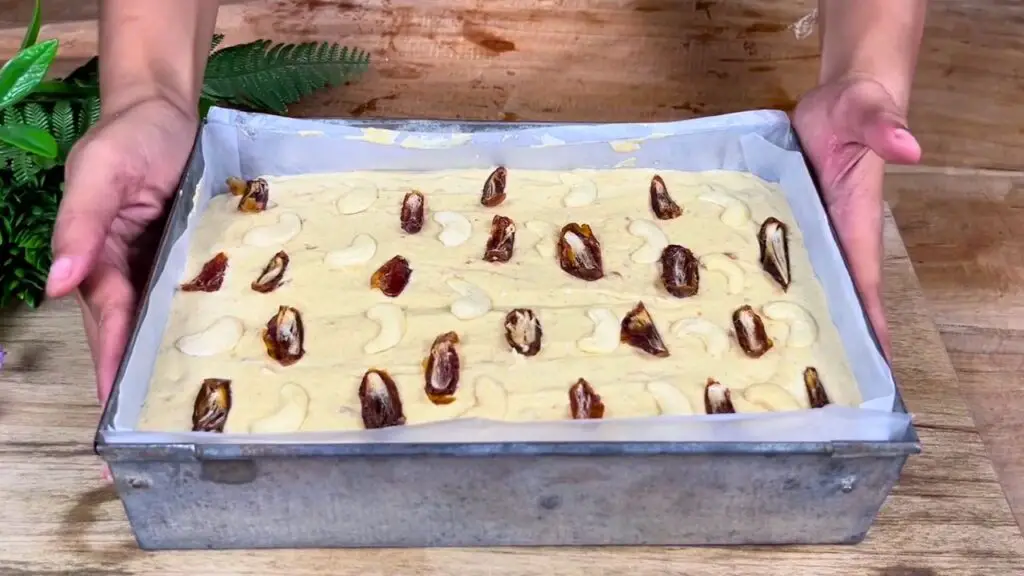 Cake batter in a lined baking pan is topped with chopped dates and cashews before baking in this best date cake recipe.