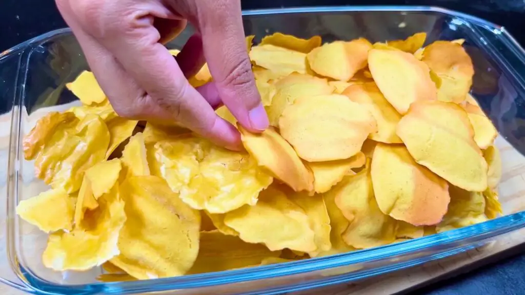 Crispy golden chickpea chips are stored in a glass container as a hand picks one up in this chickpea crisps recipe.