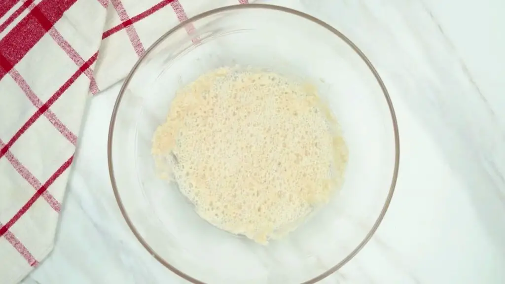 asy croissant recipe step showing a glass bowl with lukewarm water, sugar, and yeast mixture that has turned foamy and bubbly, indicating the yeast is activated.