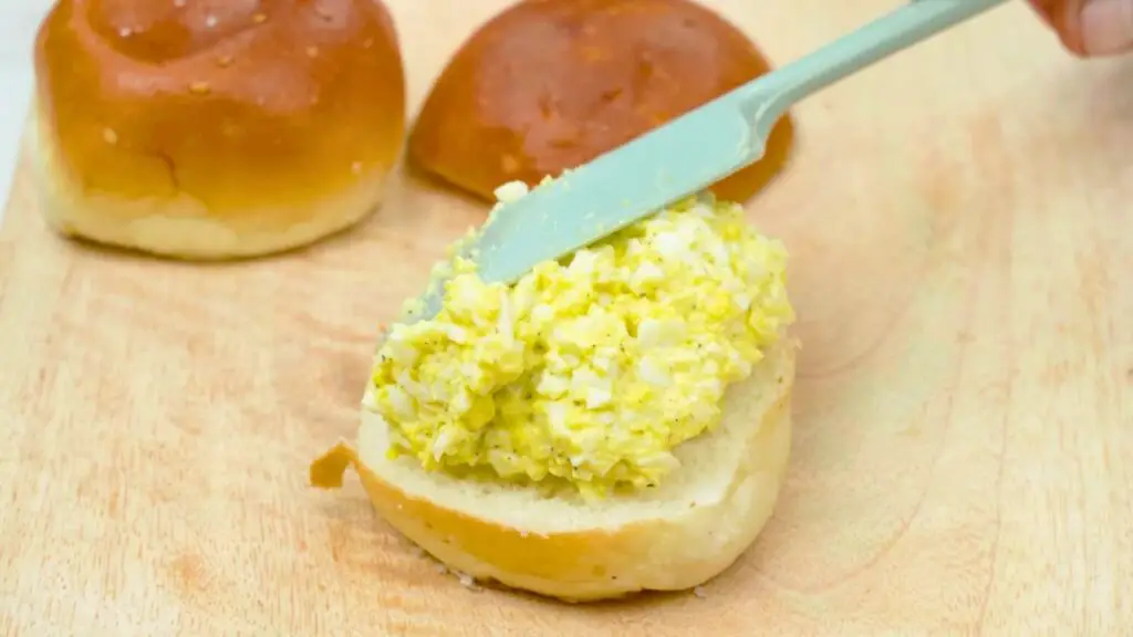 Egg salad mixture being spread onto a soft sandwich bun with a spatula on a wooden board as part of a creamy egg salad sandwich recipe