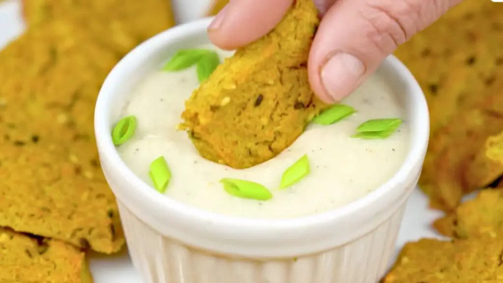Hand dipping a crispy golden cracker into a creamy dip topped with sliced green onions, served in this homemade crackers recipe.
