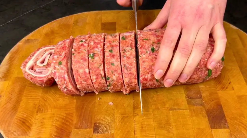 Hand slicing a Stuffed Meat Roll into even pieces on a wooden cutting board, revealing the spiral filling of ham and cheese inside.