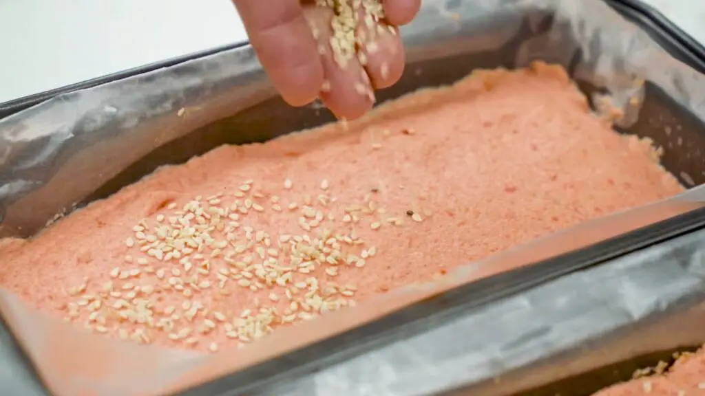 Hand sprinkling sesame seeds over red lentil batter in a parchment-lined loaf pan for the best red lentil bread recipe.