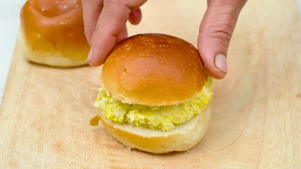 Hands placing the top bun onto a soft roll filled with egg salad as the Japanese egg salad sandwich is assembled after storage, ready to be eaten.