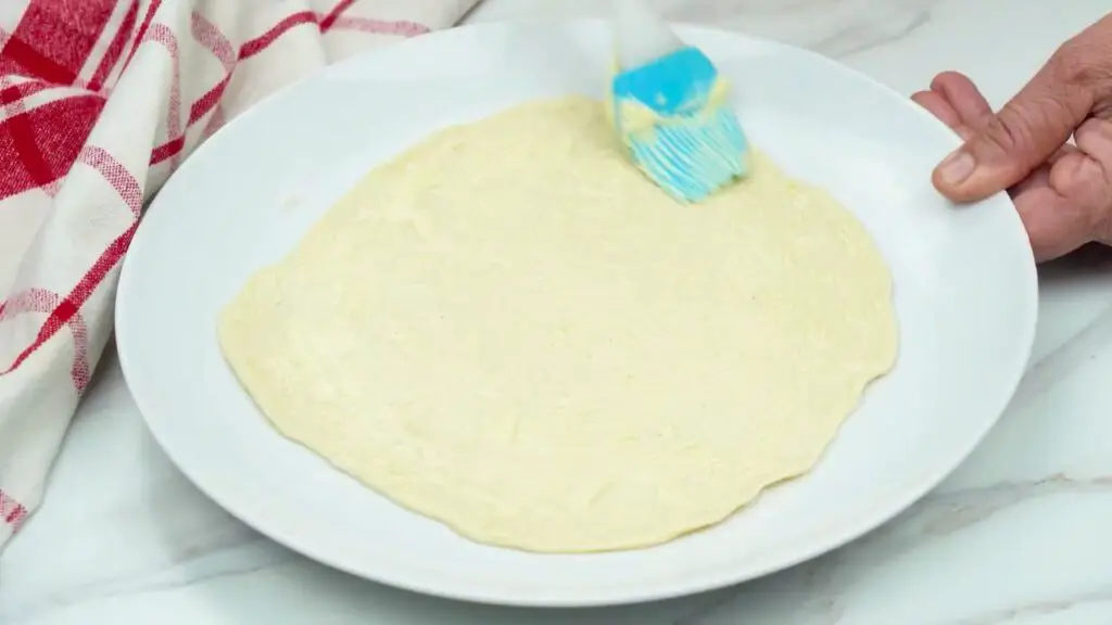 How to make butter croissants step showing flattened dough on a plate being brushed evenly with a layer of soft butter using a pastry brush, covering the surface to the edges.