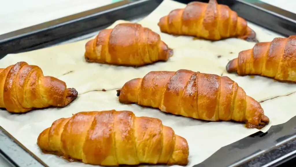 How to make croissants at home step showing freshly baked croissants on a parchment-lined baking tray, golden brown and flaky after coming out of the oven.