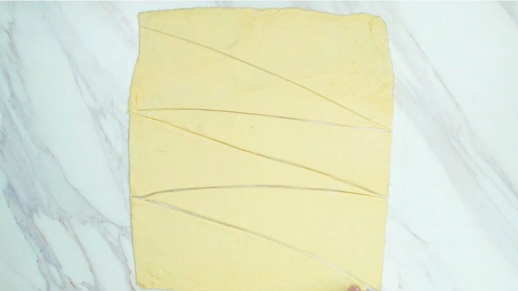 How to make croissants at home step showing rolled-out dough cut into evenly sized triangular pieces on a countertop, ready for shaping into croissants.