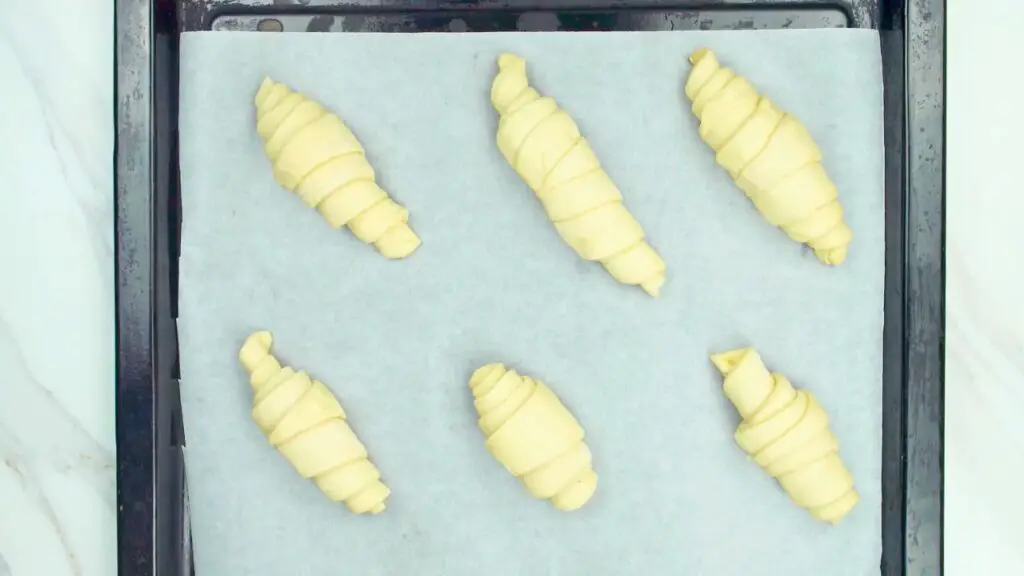How to make croissants from scratch step showing six shaped croissants placed on a parchment-lined baking tray, evenly spaced and ready for proofing or baking.