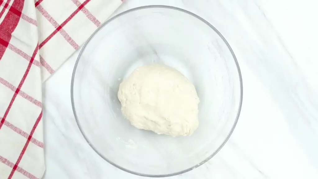 Quick and easy croissant recipe step showing a glass bowl with a rough dough formed from flour and yeast mixture, partially kneaded with no dry flour visible.