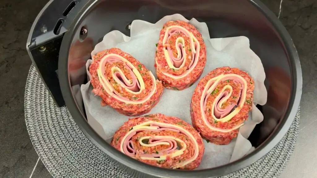 Sliced stuffed meatloaf rolls arranged on parchment paper inside an air fryer basket, ready to cook for an air fryer meatloaf.