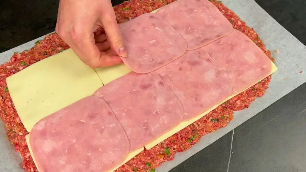 Slices of ham being layered over cheese on a flattened meat mixture, preparing the filling for a meat roll recipe.