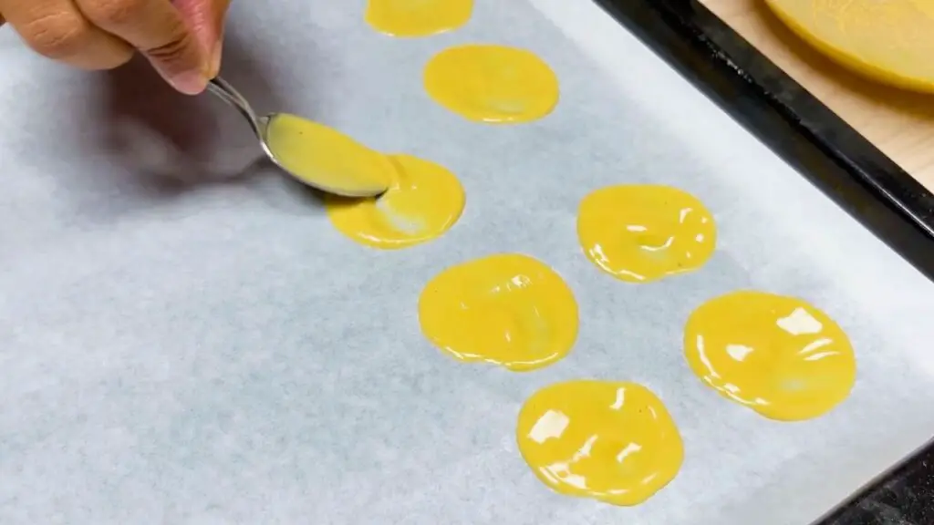 Small spoonfuls of batter are spread into thin circles on parchment paper, preparing baked chickpea chips before baking.