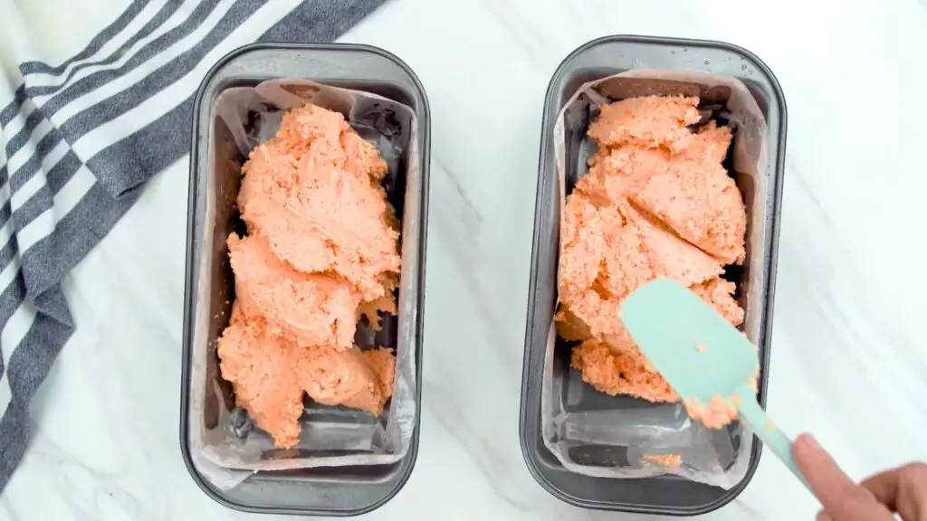 Spoonfuls of red lentil batter being divided into two parchment-lined loaf pans for lentil bread with yeast.