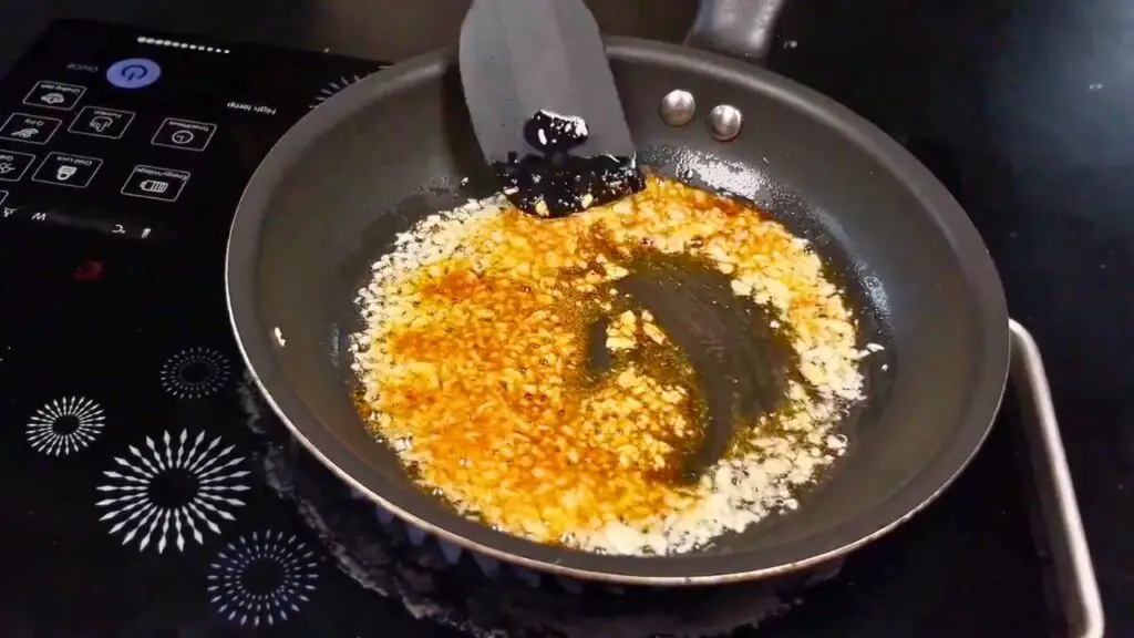 Canned sardine recipe step showing minced garlic cooking in olive oil in a frying pan, stirred until fragrant without browning.