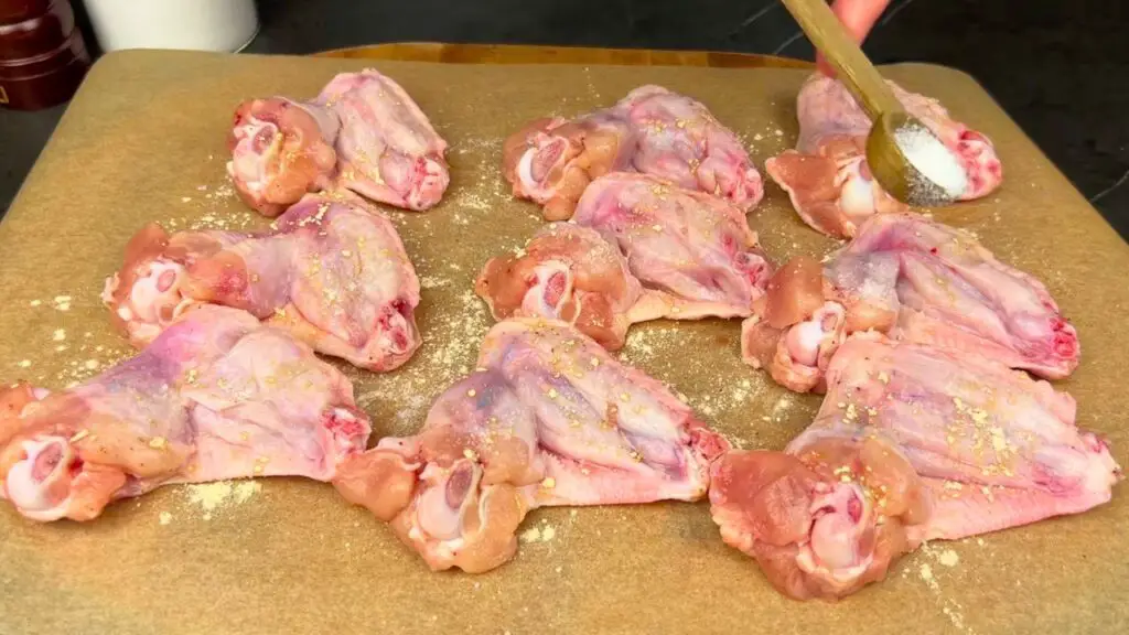 Raw chicken wings on foil-lined tray being seasoned with salt, garlic powder, and black pepper.