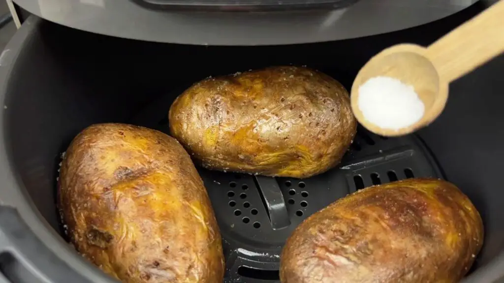 Air fryer baked potatoes recipe step showing crispy baked potatoes cooking in an air fryer basket with extra salt added before turning halfway through cooking.