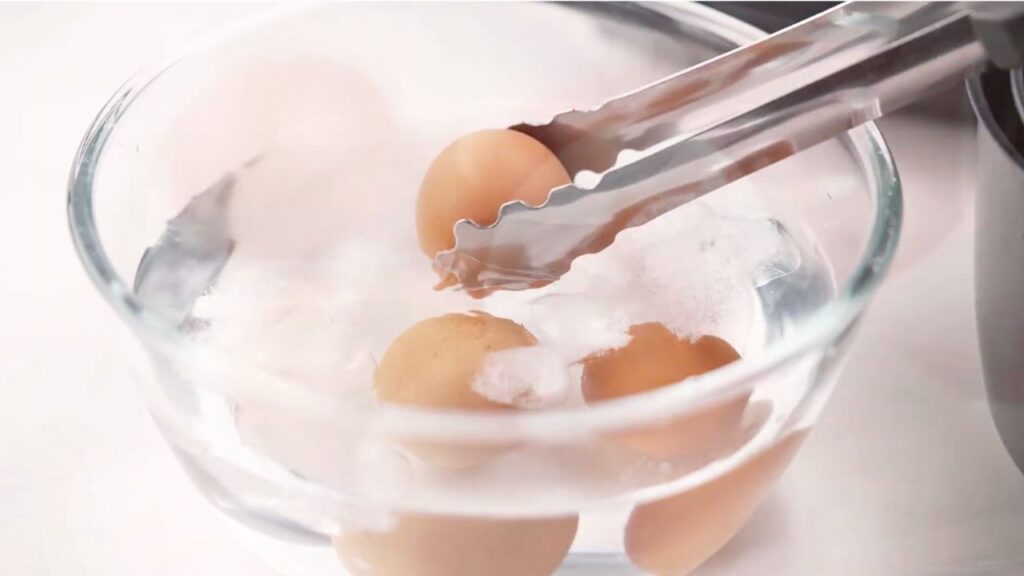 Air fryer boiled eggs being transferred to ice water with tongs to stop the cooking process and cool the eggs quickly.