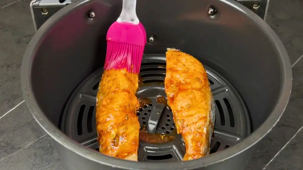 Air fryer frozen salmon being brushed with olive oil seasoning mixture after partially cooking in the air fryer basket.