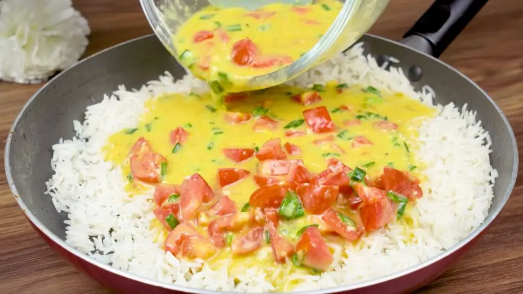 Egg mixture with tomatoes and leeks being poured over cooked rice for fried rice with eggs and vegetables.