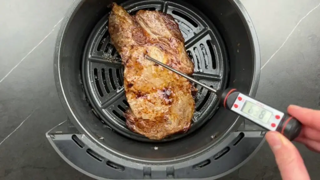 A digital meat thermometer checks the internal temperature of a cooked ribeye in the air fryer basket, showing the final doneness during this air fryer ribeye steak recipe for a well-done steak.