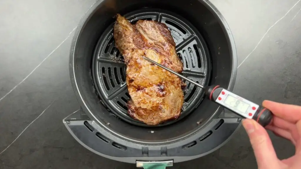 A digital meat thermometer checks the internal temperature of a cooked ribeye steak in the air fryer basket during the final stage of this easy steak air fryer recipe.