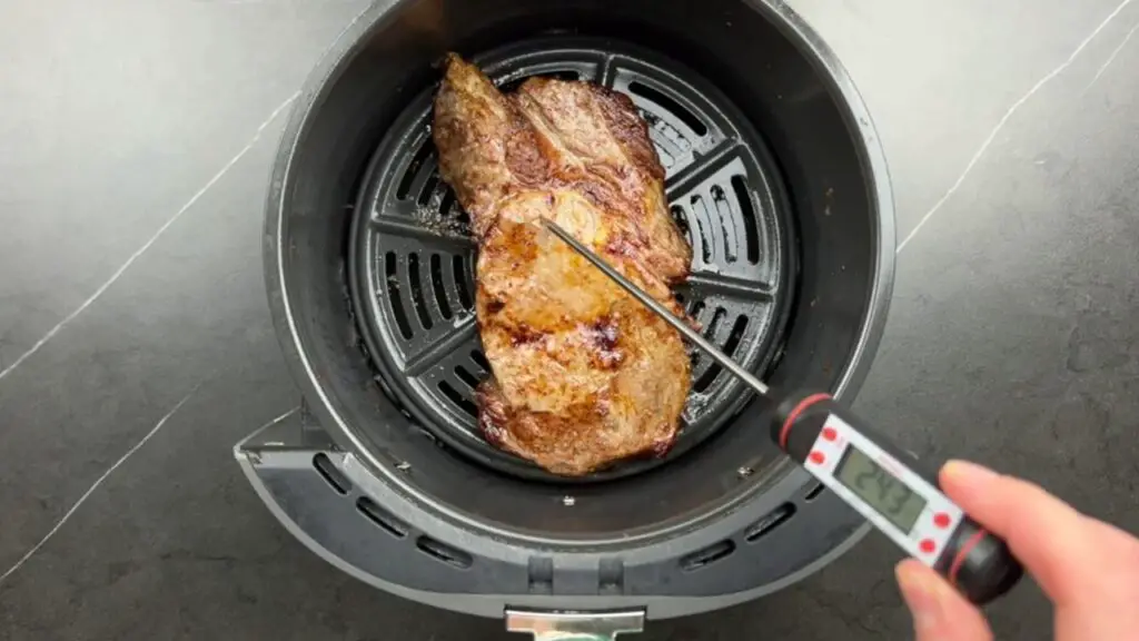 A digital thermometer checking the internal temperature of a cooked ribeye steak inside an air fryer basket, demonstrating the ideal steak in air fryer temperature for your preferred level of doneness.