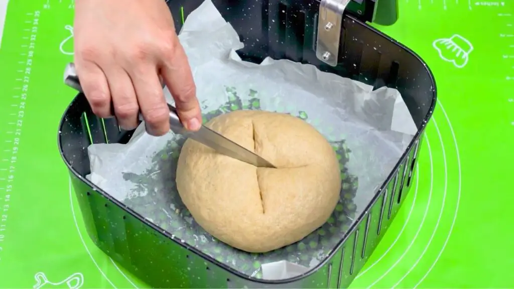 A hand scoring a round loaf of whole wheat bread in an air fryer basket, with a shallow cross cut on top before baking.