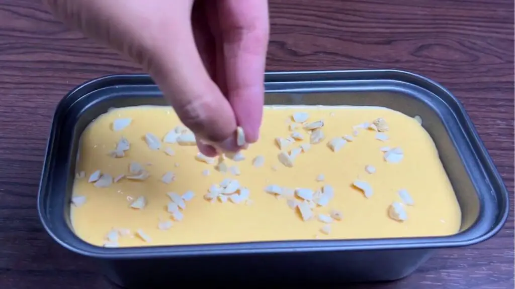 Chopped nuts being sprinkled over a creamy mango ice cream mixture in a loaf pan, adding texture and toppings before freezing the homemade mango ice cream.
