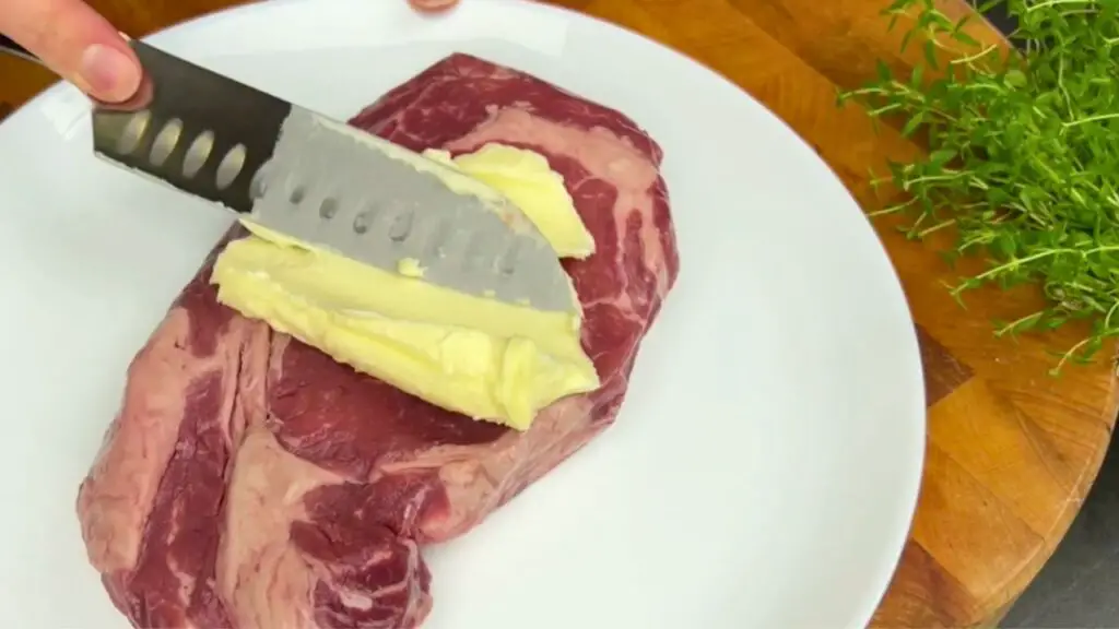 Cold ribeye steak on a white plate with softened butter being spread across the top using a knife, preparing the steak for an easy air fryer steak recipe.