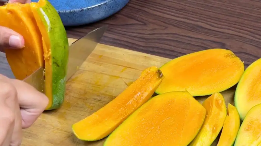Fresh ripe mango being sliced on a wooden cutting board, with bright orange mango slices arranged nearby for making homemade mango ice cream.