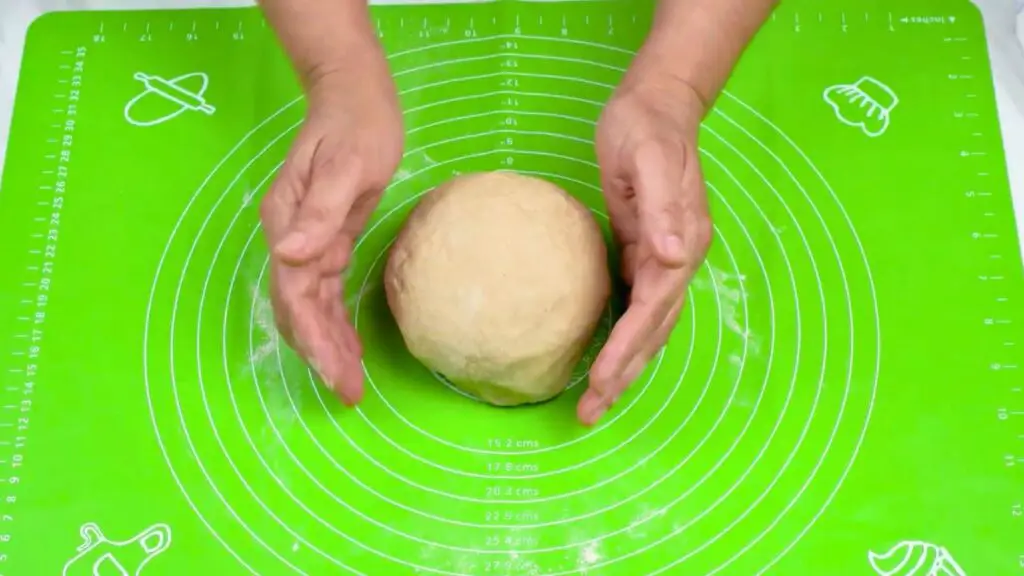 Hands shaping a smooth ball of whole wheat bread dough on a lightly floured green baking mat.